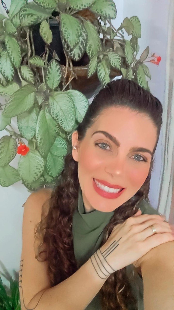 Flávia Maitri sorrindo perto de uma planta, em ambiente doméstico